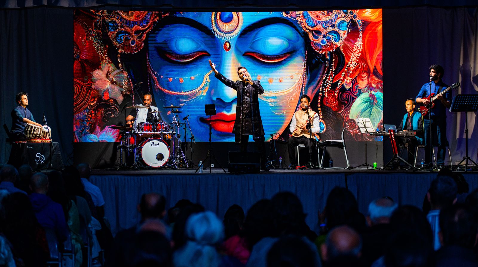 The full band on stage, tabla, drums, vocals, bansuri, keys and guitar, beneath a vast artwork of the Goddess