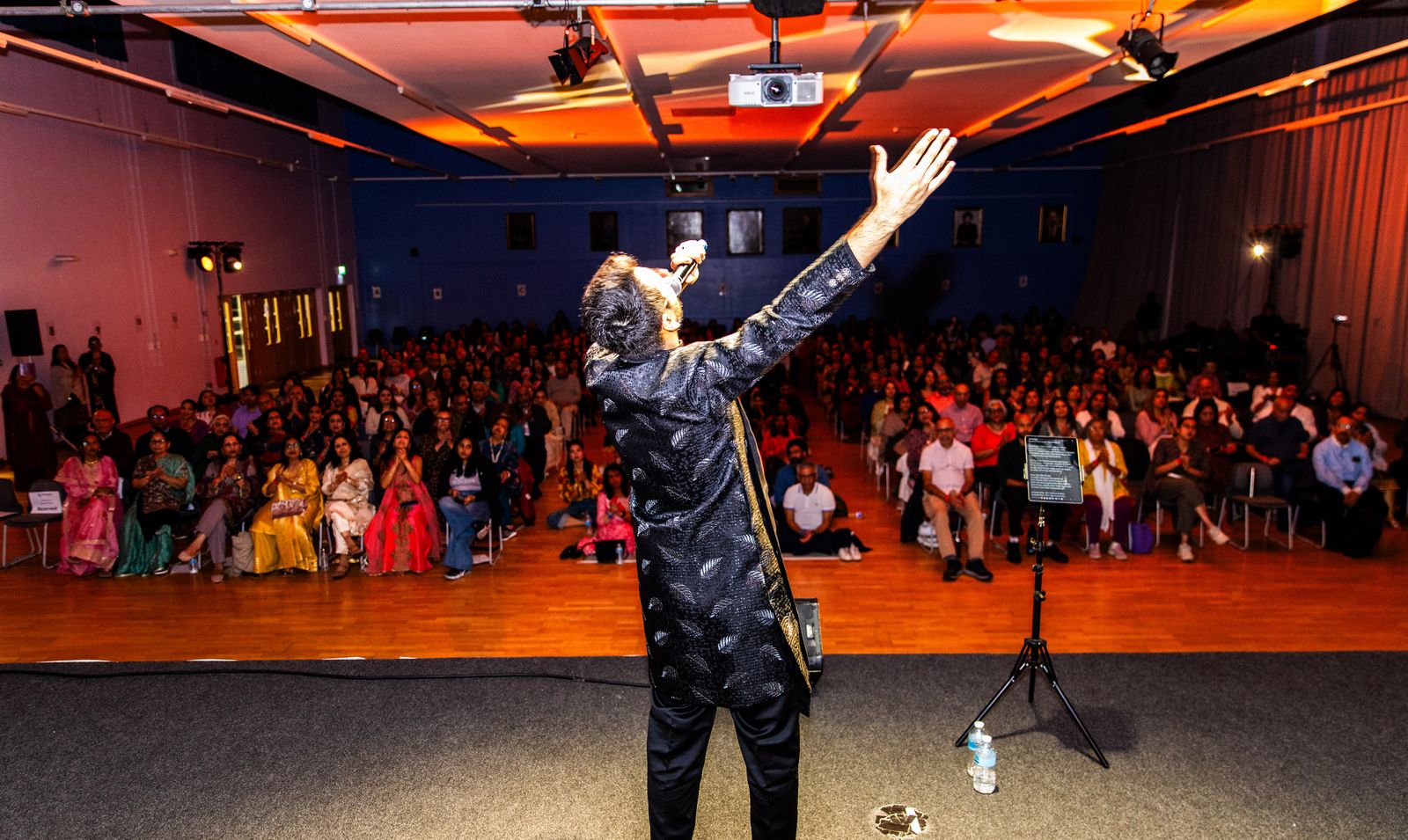 Priyesh Shah on stage with his arm raised, facing a packed hall of families clapping along