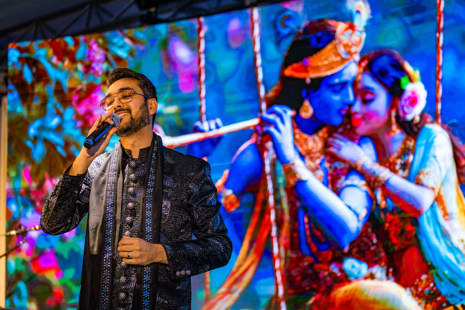 Priyesh Shah singing with eyes closed in front of a screen showing Radha and Krishna on a swing