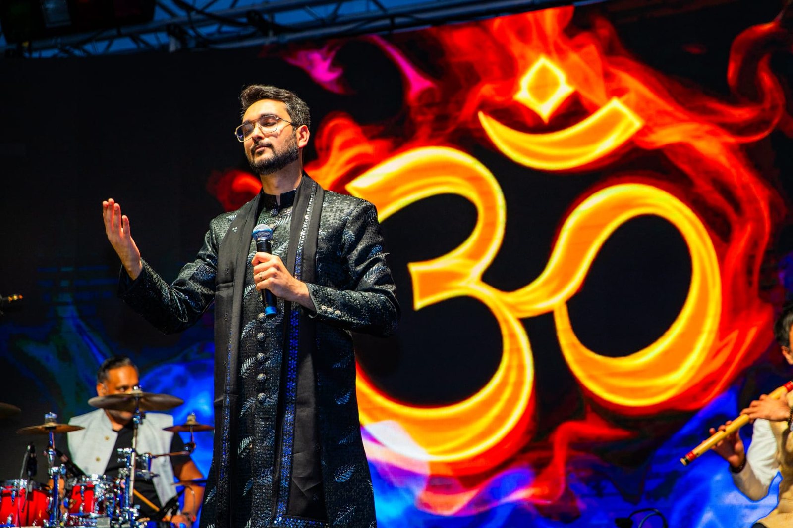 Priyesh Shah singing with eyes closed in front of a blazing Om on the stage screen