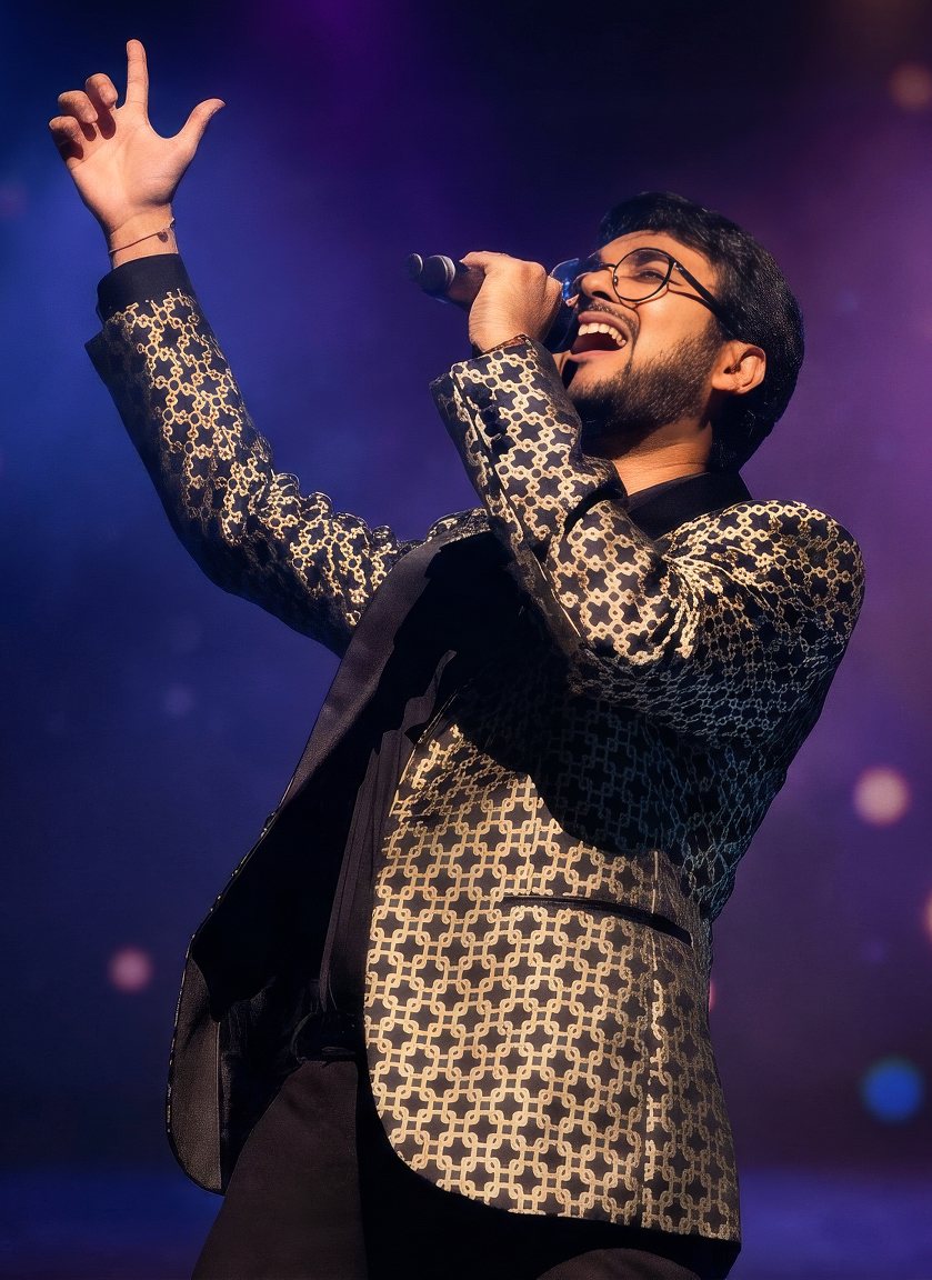 Priyesh Shah singing on stage, one arm raised, in a gold patterned jacket